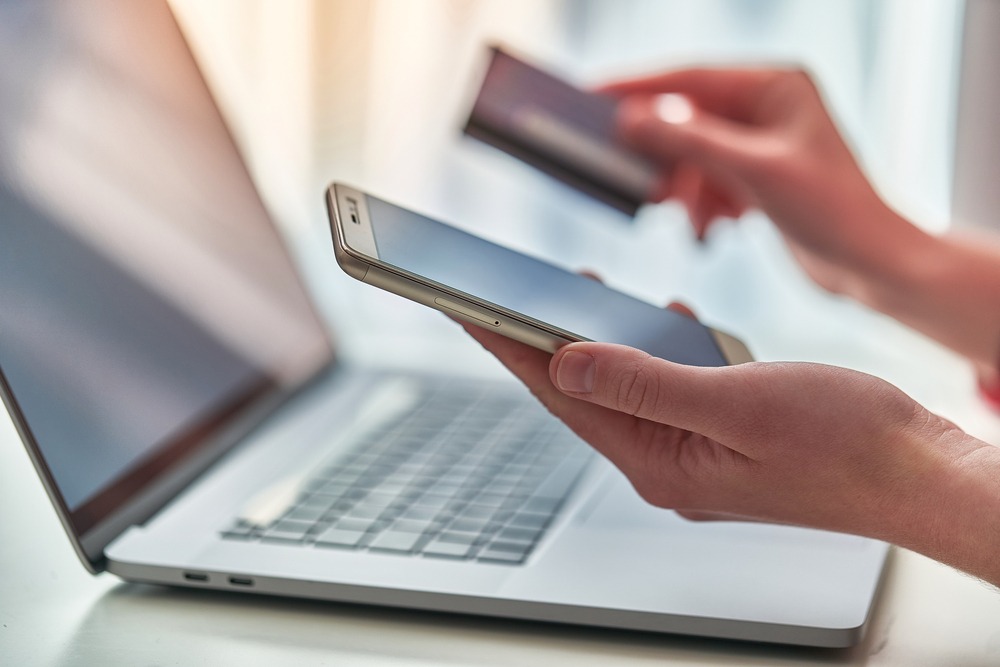 Closeup of a laptop and two hands, one hand holds a phone and the other hand a bank card