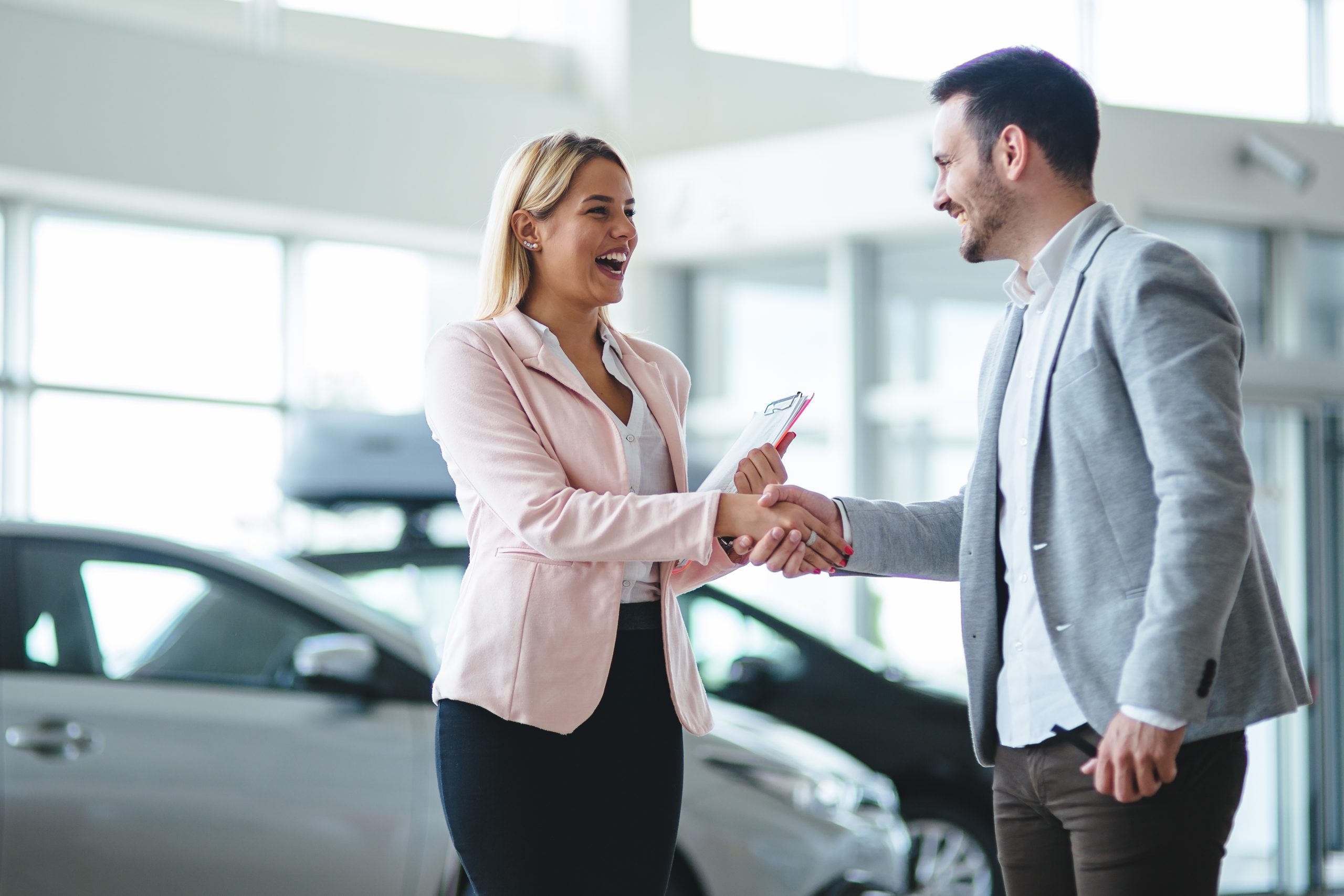 Man and woman shaking hands at a dealership and the woman holds paperwork in her other hand