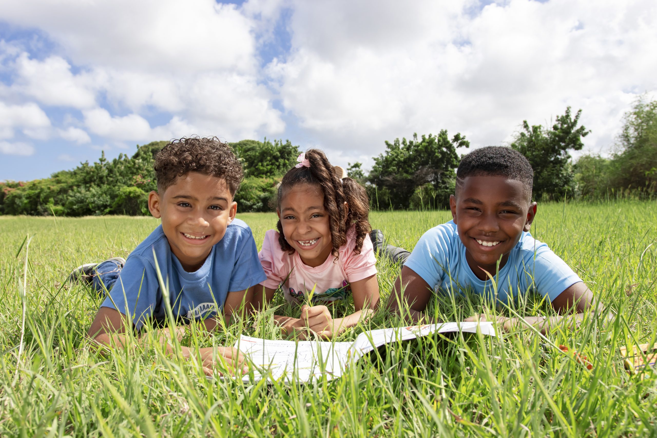 Close up of three children looking in the camera while reading something. The children are laying next to each other in the grass on their belly.