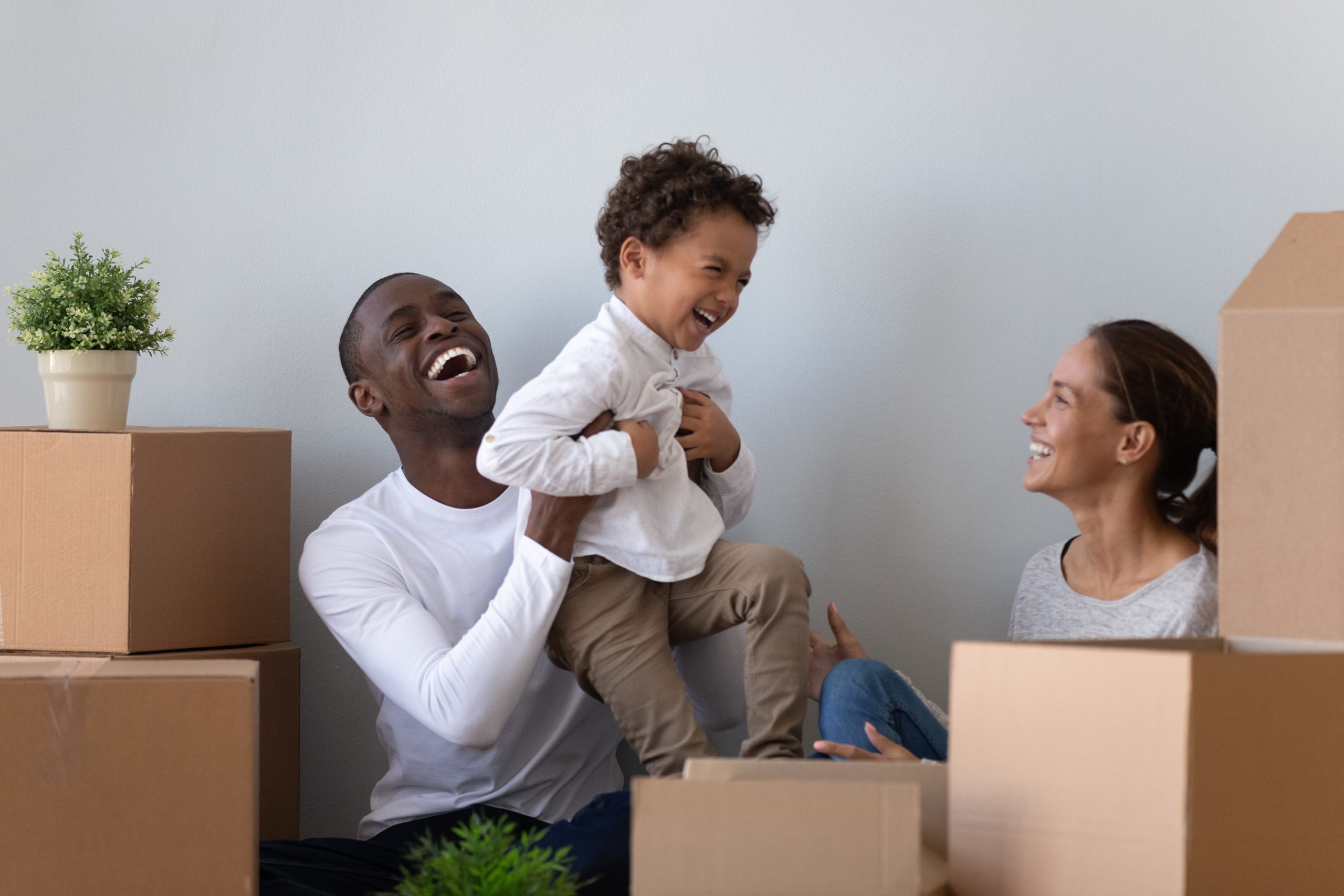 Man, woman and son are having fun in between a couple of boxes and some plants. The man is holding the son in his hands.