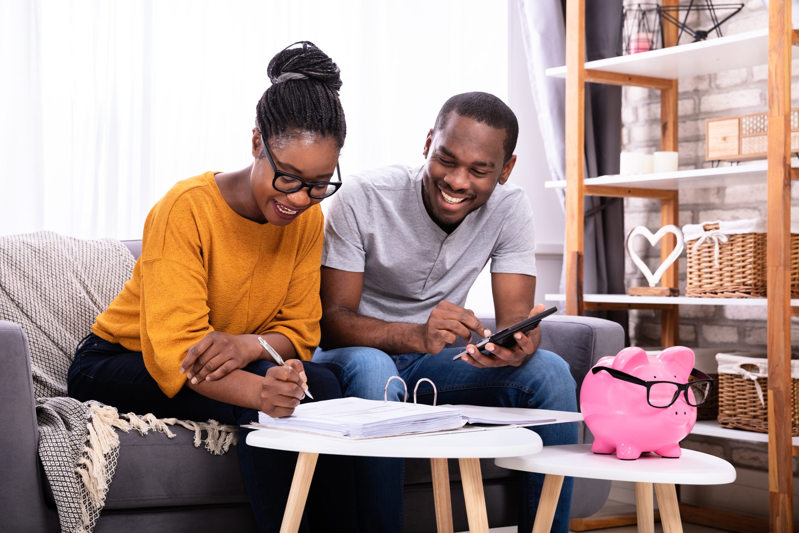 A couple on the couch. The man is holding a calculator and the woman is writing in a folder. On the table is a piggy bank.